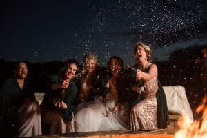 Bride and bridesmaids open champagne by the fire pit